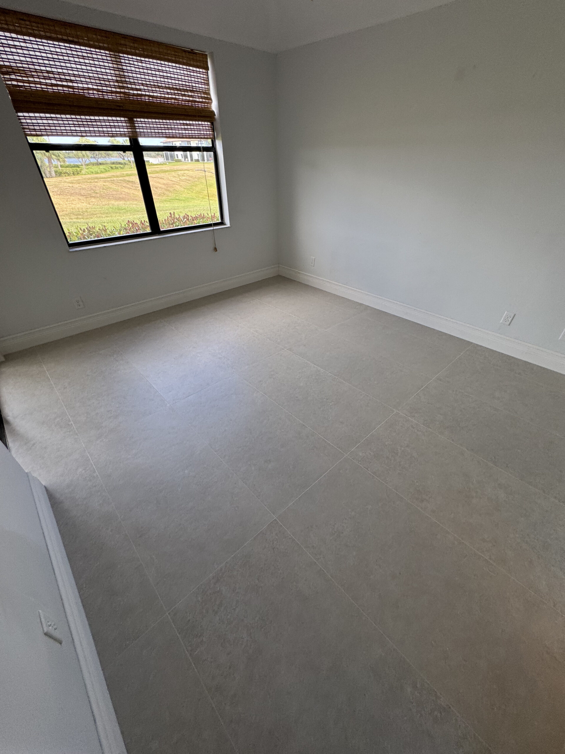 An empty room with light-colored tiled flooring, pale walls, and a large window with brown woven blinds partially open, letting in natural light.