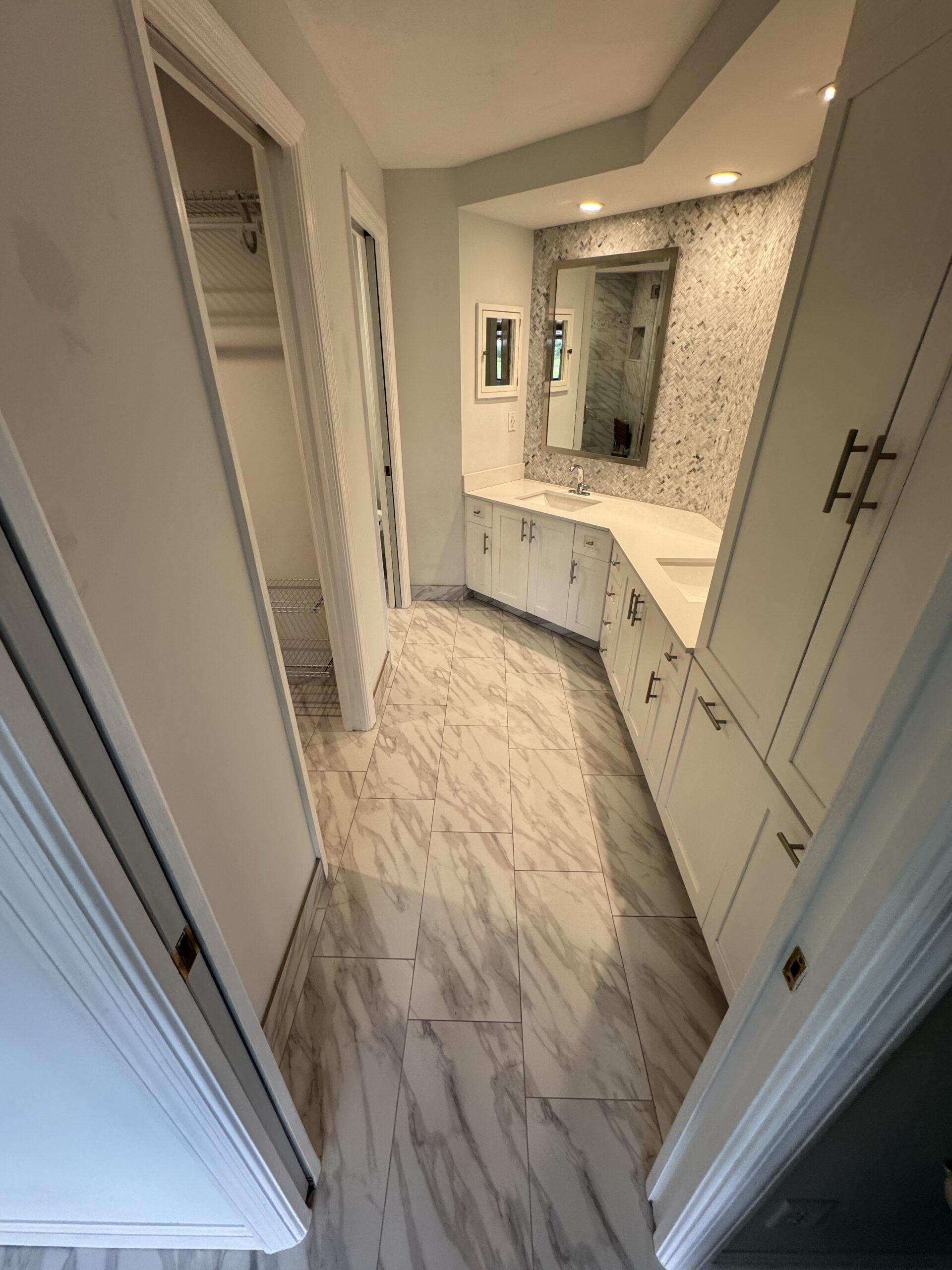 A modern bathroom with marble-patterned tile flooring, white cabinets, a large mirror, and recessed lighting. There is an open closet with wire shelving visible through a doorway on the left.