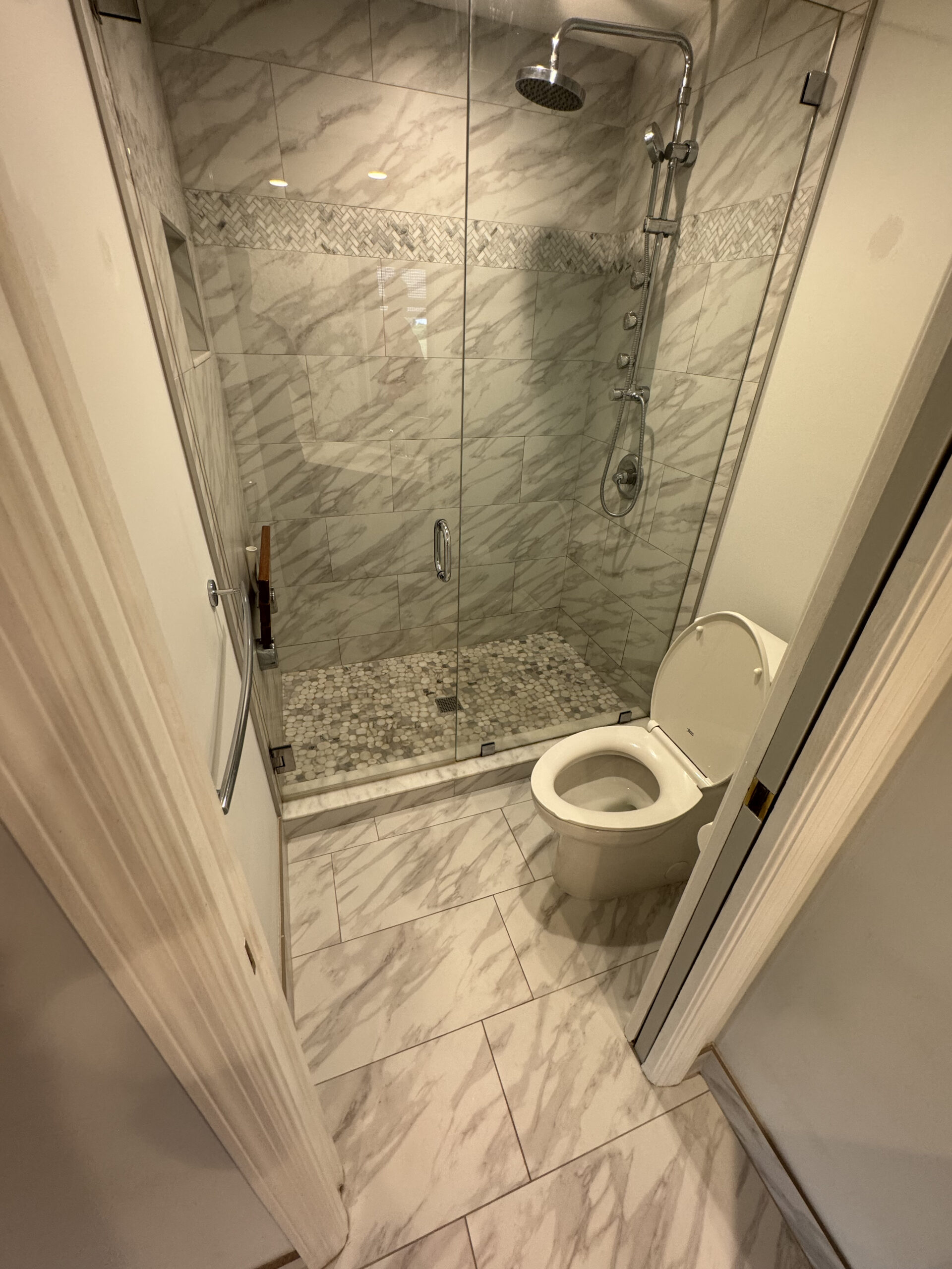 A bathroom with marble-patterned tile, a glass-enclosed shower with silver fixtures, and a white toilet next to the shower. The shower has a tiled floor and decorative tile accent on the wall.